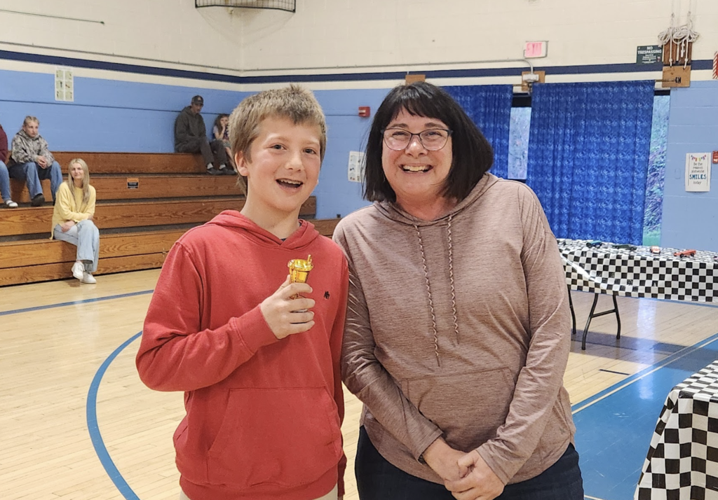 A student in a red sweatshirt holds a small gold trophy while standing next to a smiling staff member in a gym.