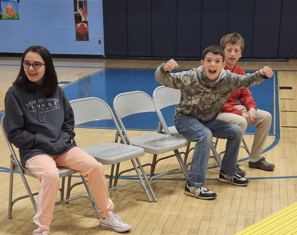 Three students sit on folding chairs in a gym, reacting excitedly to a race. One student leans forward with arms raised in celebration while others watch.