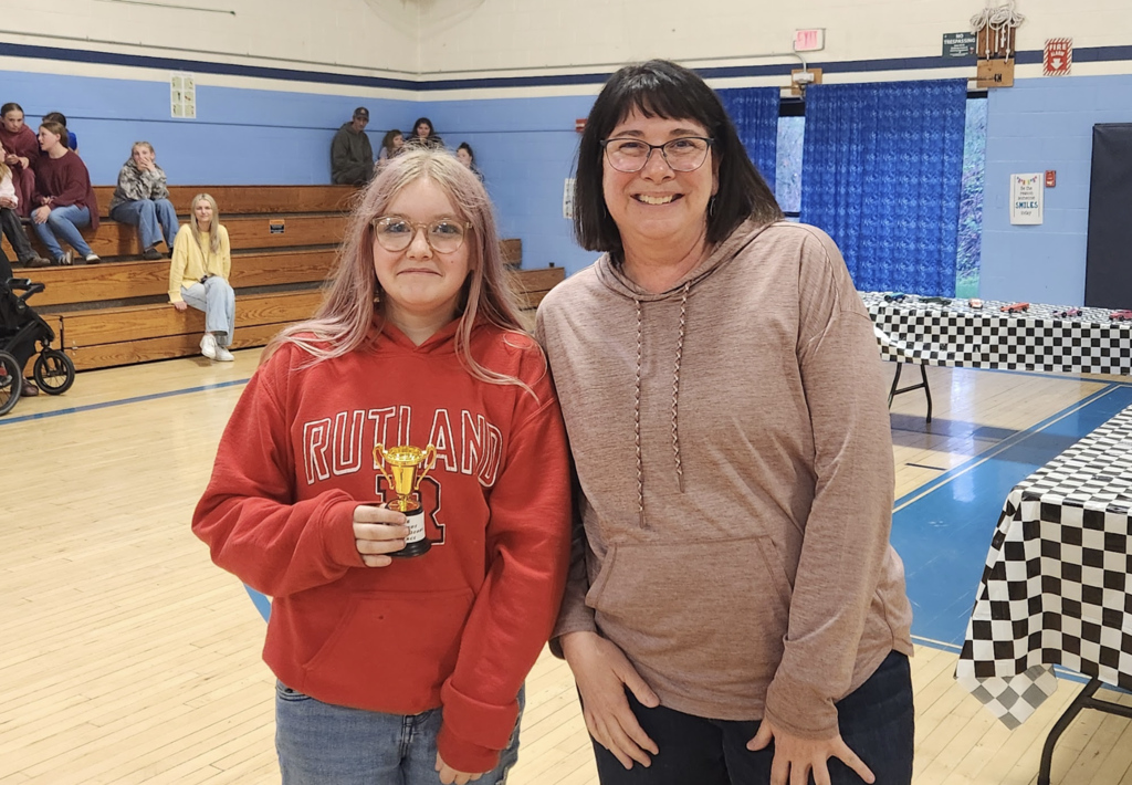 A student wearing a red hoodie and glasses holds a gold trophy while standing next to a staff member, both smiling in a gym.