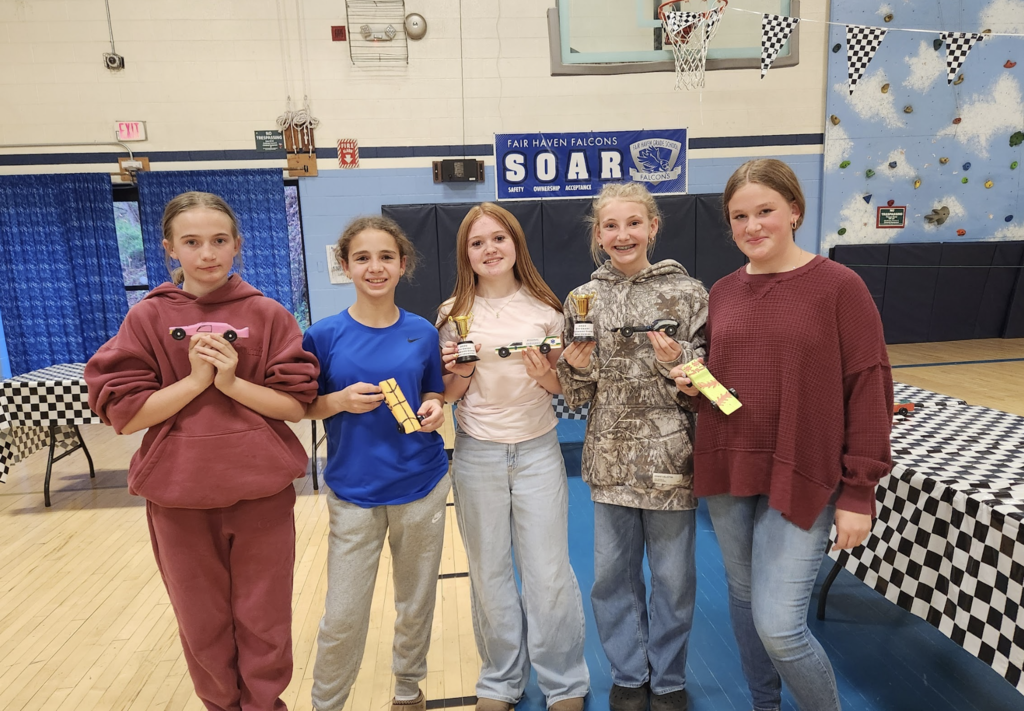 Five students stand together in a gym holding their pinewood derby cars and small trophies, smiling proudly after the race.