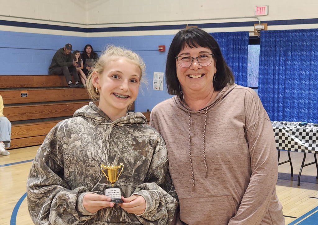 A student holding a trophy stands next to a staff member in a gym, both smiling for the camera with pinewood derby tables in the background.