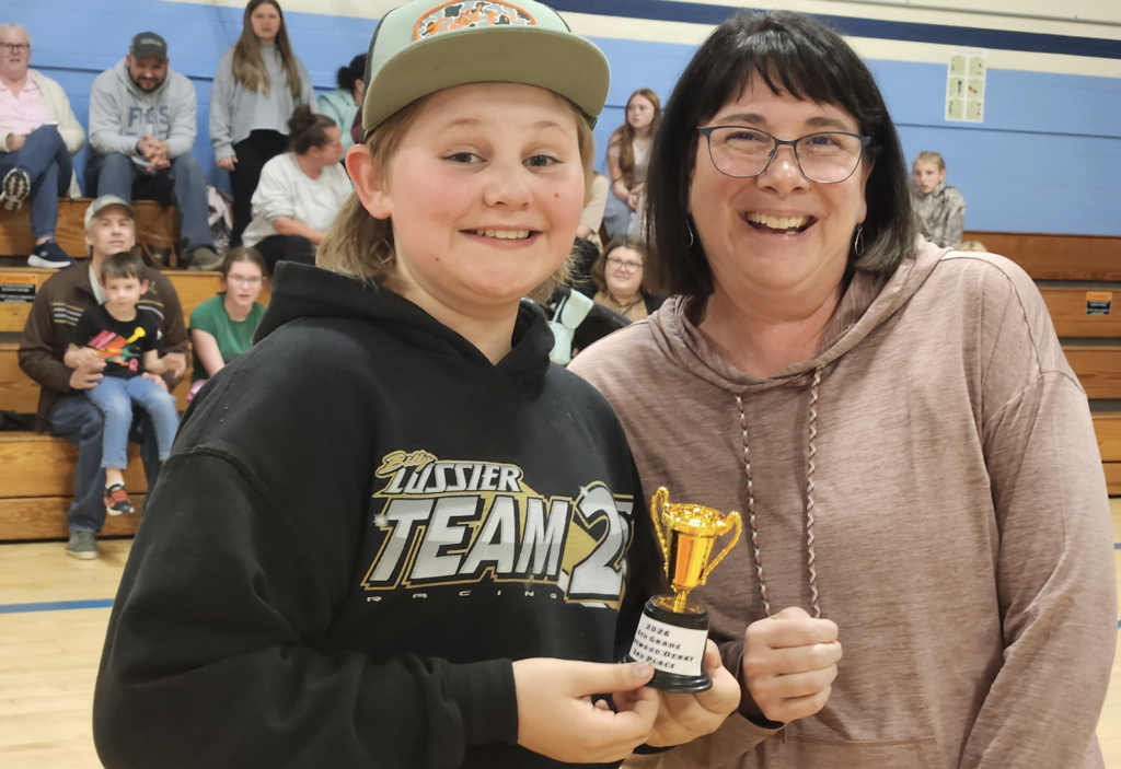 A student and a staff member smile while holding a small gold trophy together in a gymnasium, with other students seated on bleachers behind them.