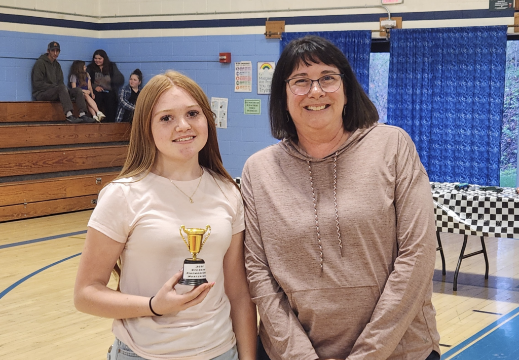 A student holding a trophy stands next to a staff member in a gym, both smiling for the camera with pinewood derby tables in the background.