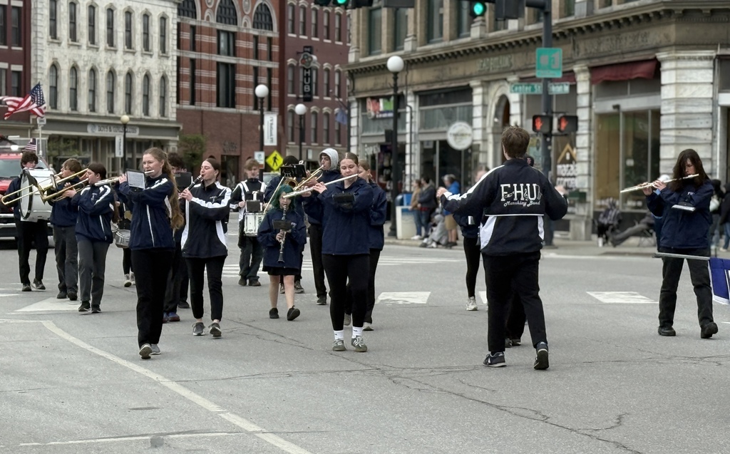 The FHUMHS marching band performs while marching through a downtown street, with students playing brass and woodwind instruments and a conductor leading in front of them.