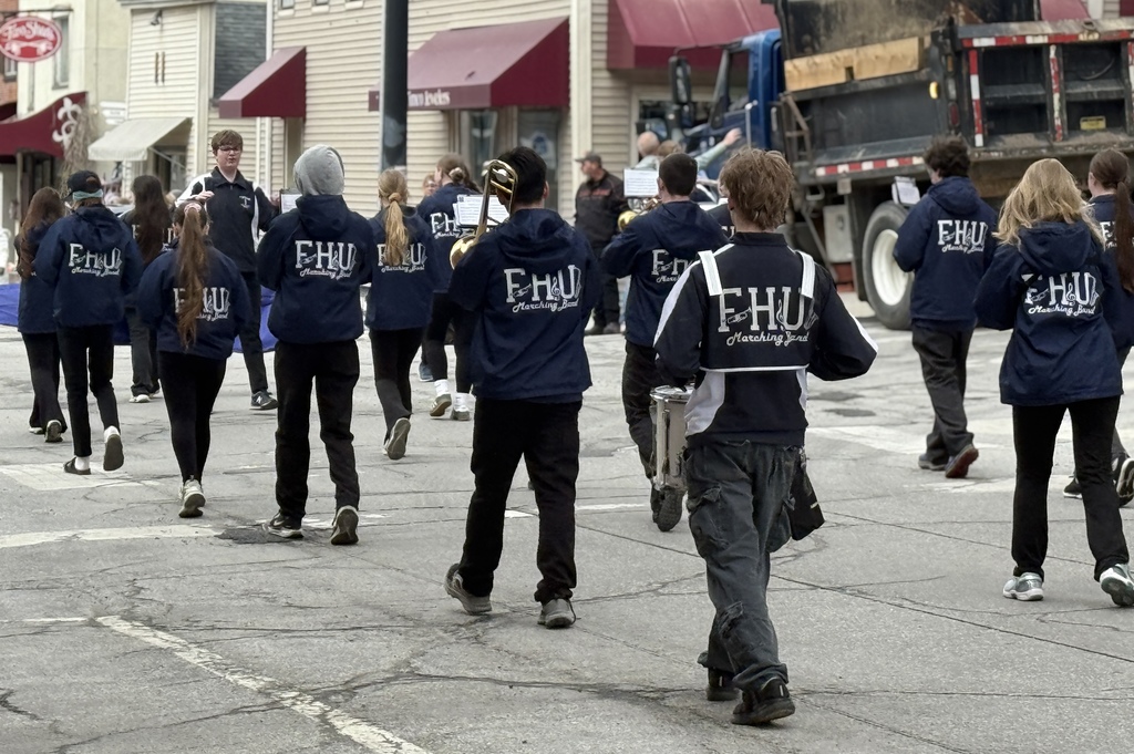 Members of the FHUMHS marching band, wearing navy jackets with “FHU Marching Band” on the back, walk down a street in formation during a parade, carrying instruments.