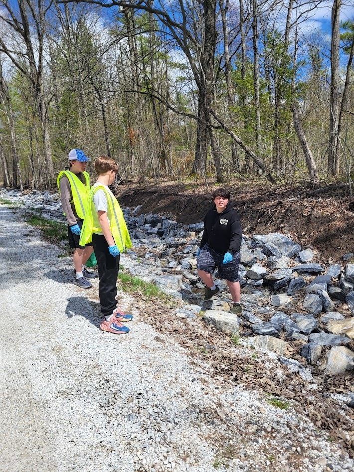 Three students wearing safety vests and gloves stand along a gravel roadside and rocky ditch, picking up litter on a sunny day.