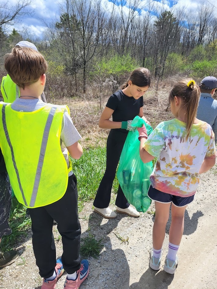 A group of students in gloves and casual clothes gather along a roadside, placing trash into a large green bag during a cleanup.