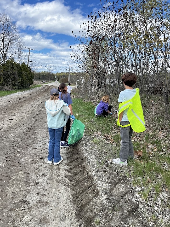 Students stand along the edge of a dirt road and grassy embankment, picking up trash near bushes and placing it into a green bag.