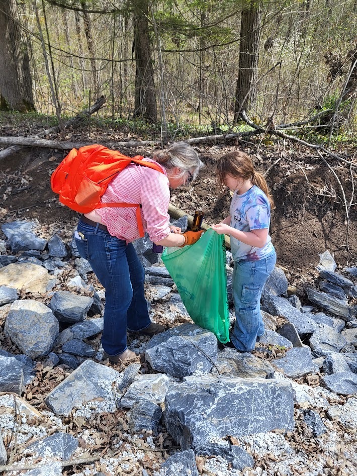 An adult with an orange backpack and a student stand on rocks near a ditch, placing collected trash into a large green bag.