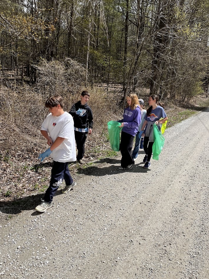 A small group of students walks along a gravel road, scanning the ground and collecting litter into green bags near a wooded area.