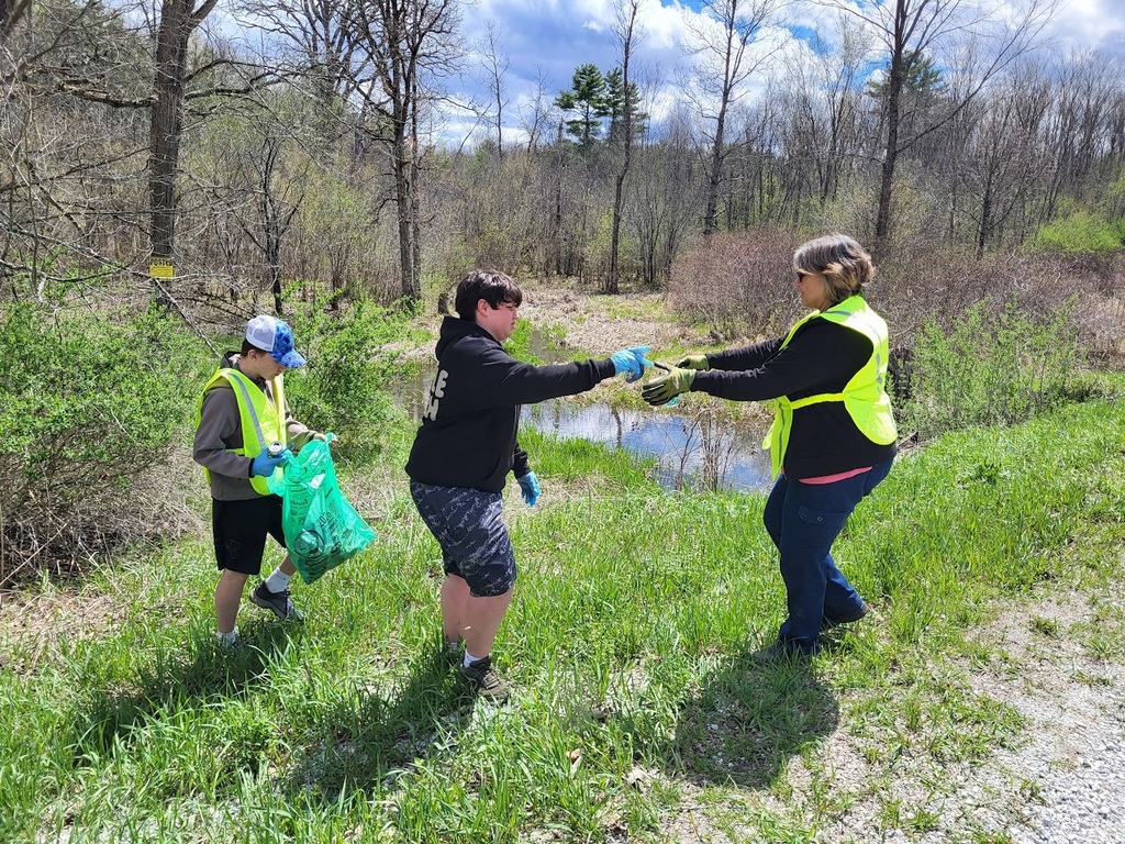 Two students in gloves pass a piece of trash to an adult wearing a safety vest beside a small stream, while another student holds a full green trash bag.