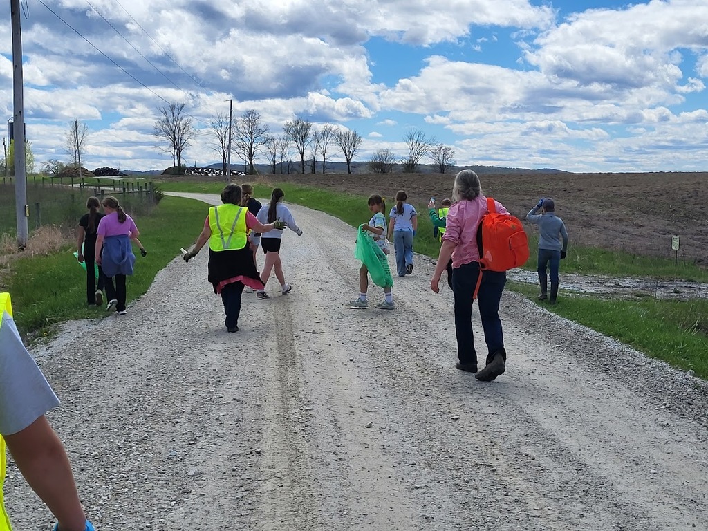 Students walk down a country road with green trash bags, continuing their roadside cleanup on a partly cloudy day.