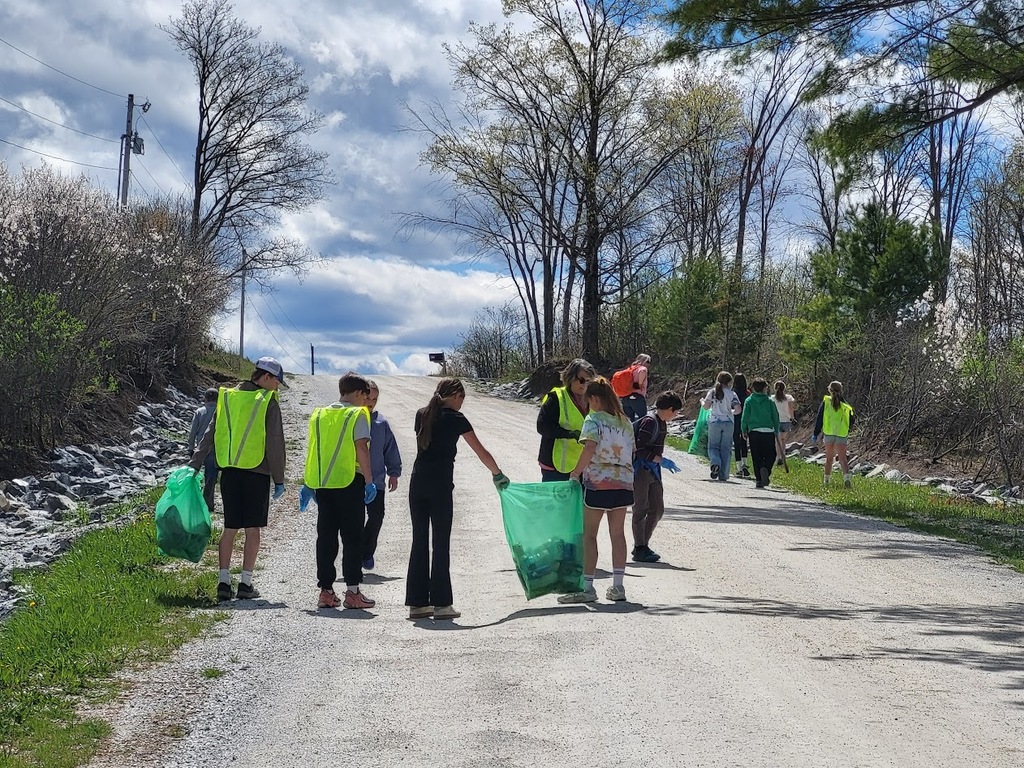 Students wearing safety vests walk along a gravel road carrying green trash bags, collecting litter under a bright sky.
