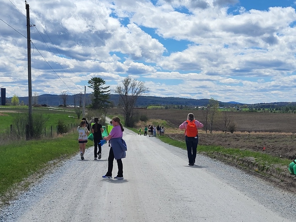 A group of students and an adult spread out along a rural dirt road, picking up trash with green bags, with open fields and hills in the background.