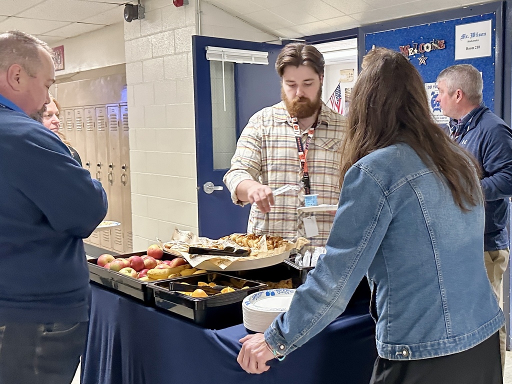 A staff member serves himself from a tray of pastries while others stand nearby holding plates in a school hallway.