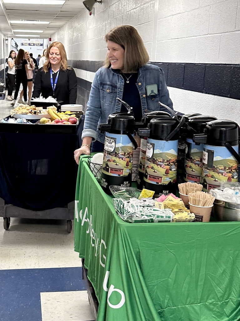 A woman smiles while pushing a cart with coffee containers down a school hallway, followed by another staff member pushing a cart with fruit and pastries.