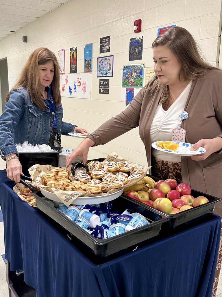 Two staff members serve themselves breakfast items from a table while others stand nearby in the hallway.