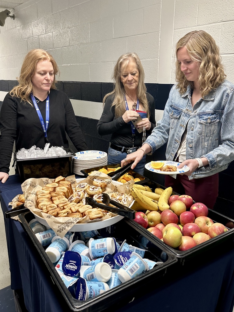Three staff members stand behind a table filled with breakfast items including apples, bananas, yogurt, and pastries as one serves herself.