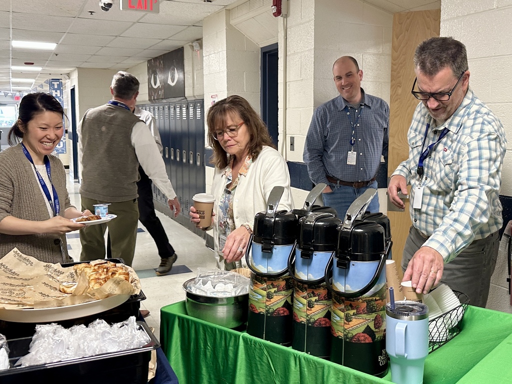 Several staff members gather around a coffee station in a hallway, pouring drinks and selecting breakfast items while chatting.