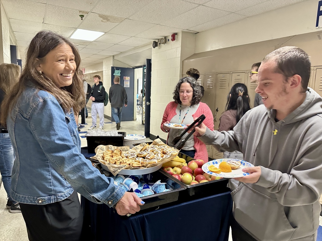 A smiling staff member hands food to another while standing beside a table filled with pastries, fruit, and yogurt.