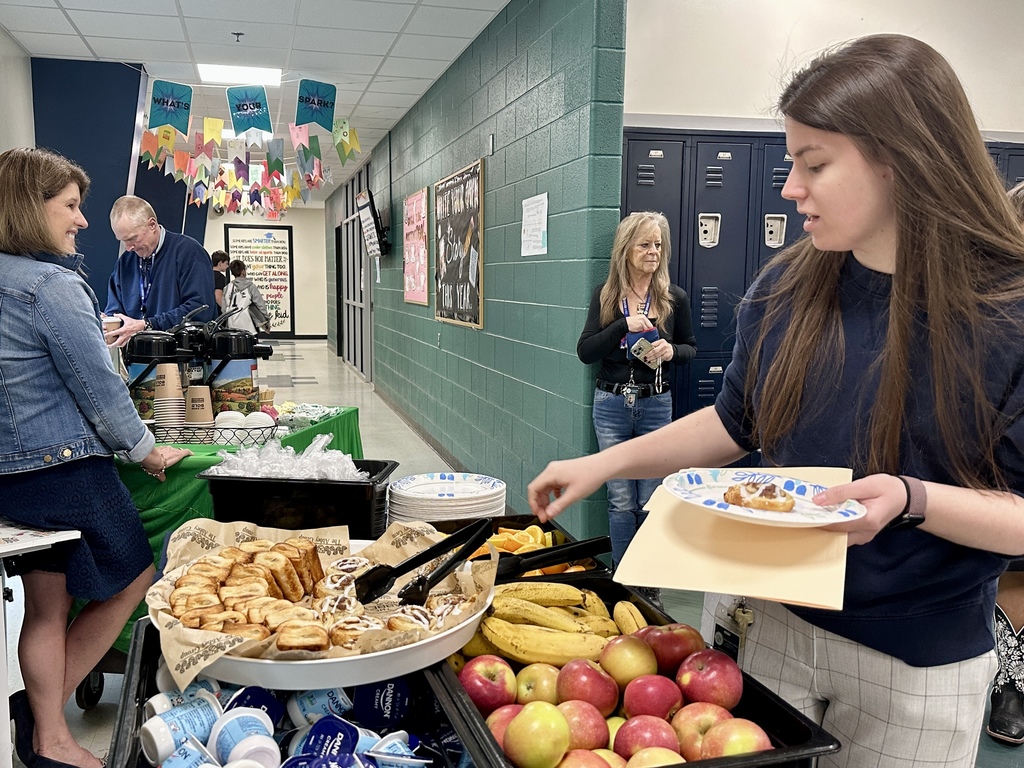 A staff member selects food from a table filled with pastries, fruit, and yogurt while holding a plate.