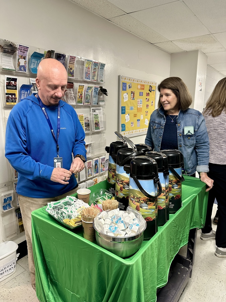 A man and woman stand behind a green-covered table in a school hallway preparing coffee and tea, with creamers, stir sticks, and cups arranged neatly.