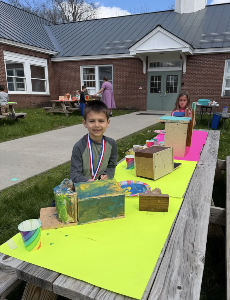 A smiling young student wearing a medal sits at a picnic table with a neon yellow surface, proudly displaying a painted wooden birdhouse. Other students work in the background near the school building.