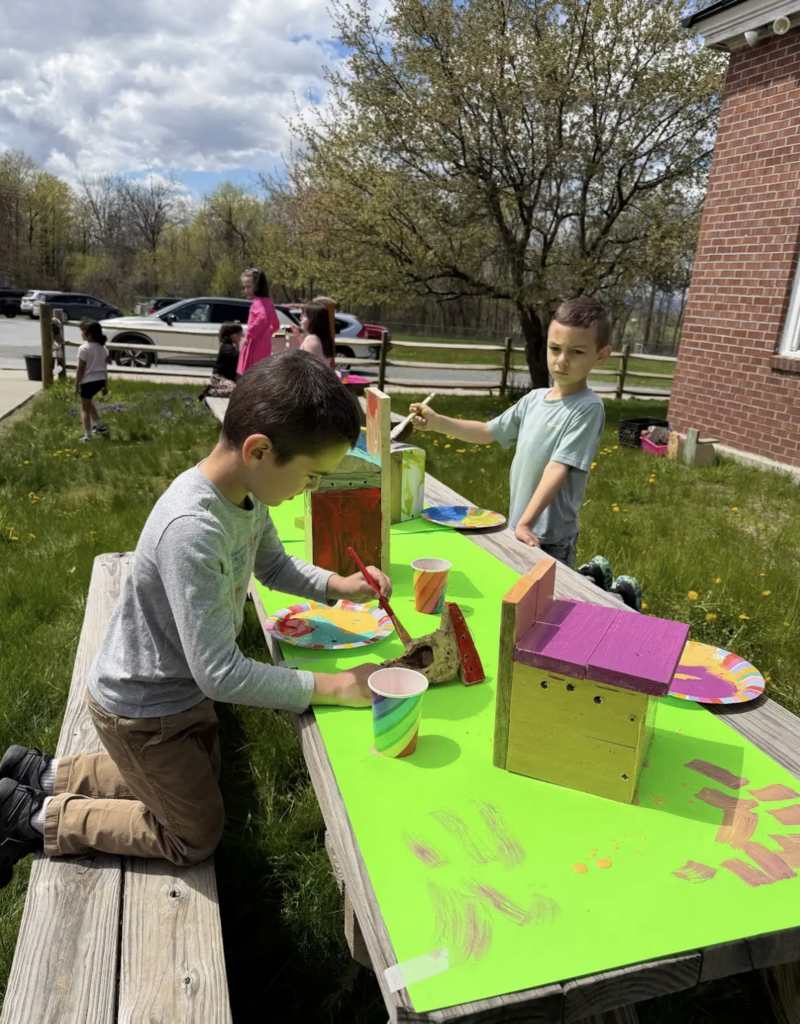 Two young students paint birdhouses at a bright green-covered picnic table outdoors. One child kneels while painting carefully, and another stands holding a paintbrush. Grass, trees, and parked cars are visible in the background under a partly cloudy sky.