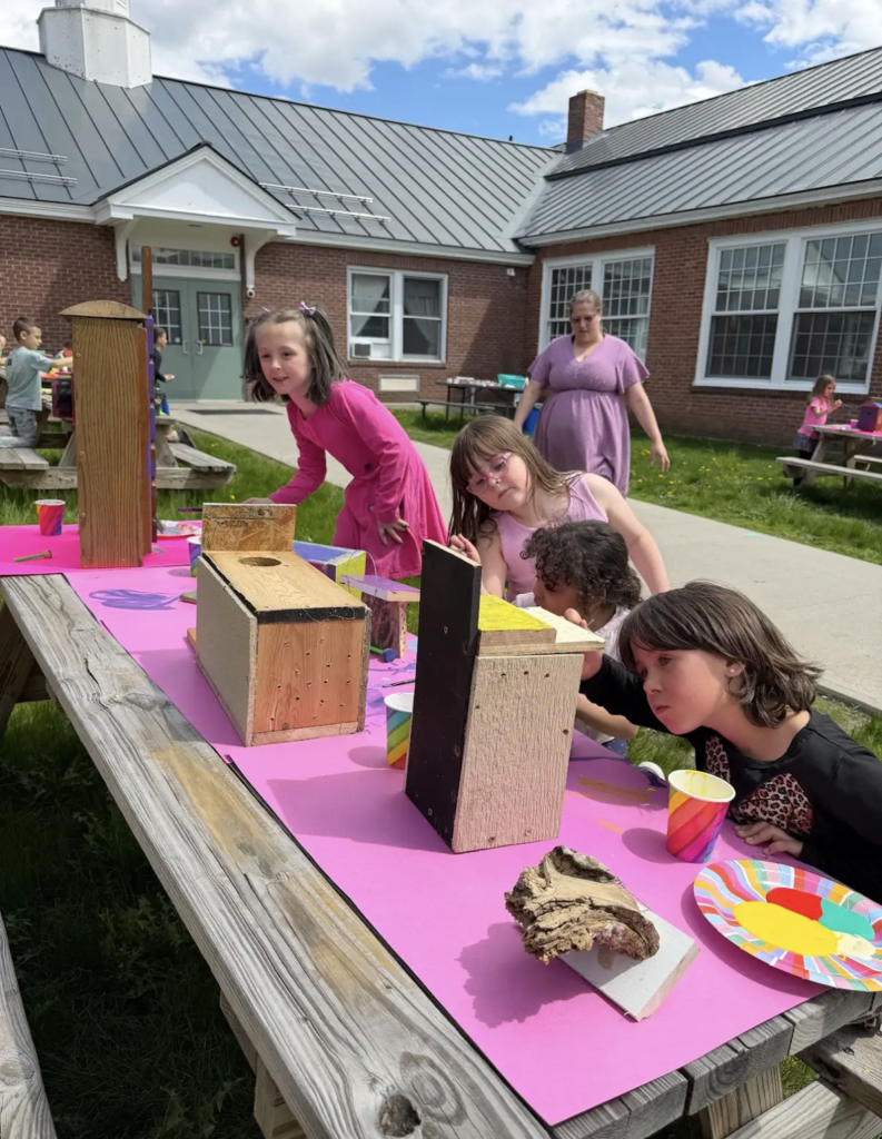 A group of first-grade students gathers around a pink-covered picnic table outside, carefully painting wooden birdhouses. One student leans in closely to add detail while others watch and work. A teacher stands nearby, supervising. The brick school building and grassy area are visible behind them.