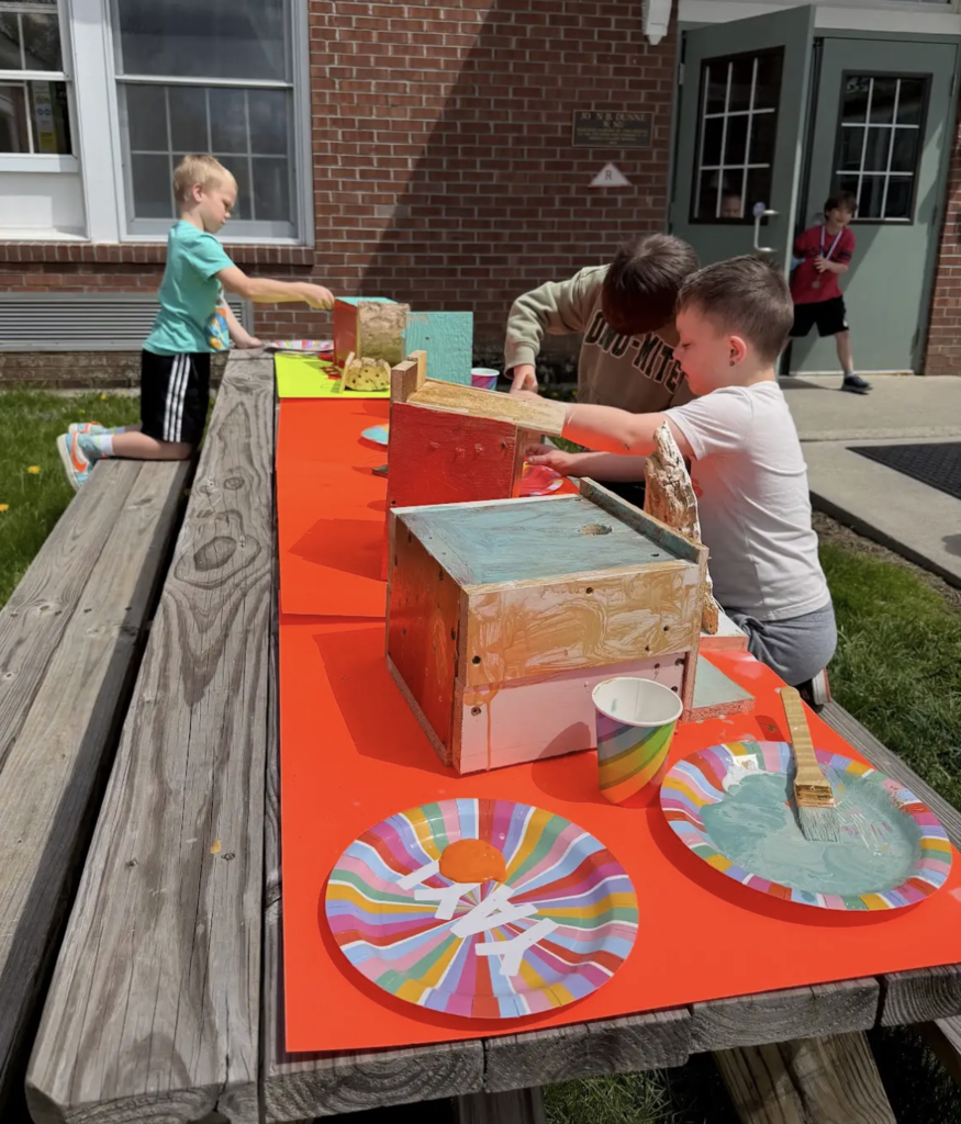 Three young students sit and kneel at a long outdoor picnic table covered with a bright orange tablecloth, painting small wooden birdhouses and bee homes. Paint plates, brushes, and cups are spread across the table. A school building with brick walls is in the background.