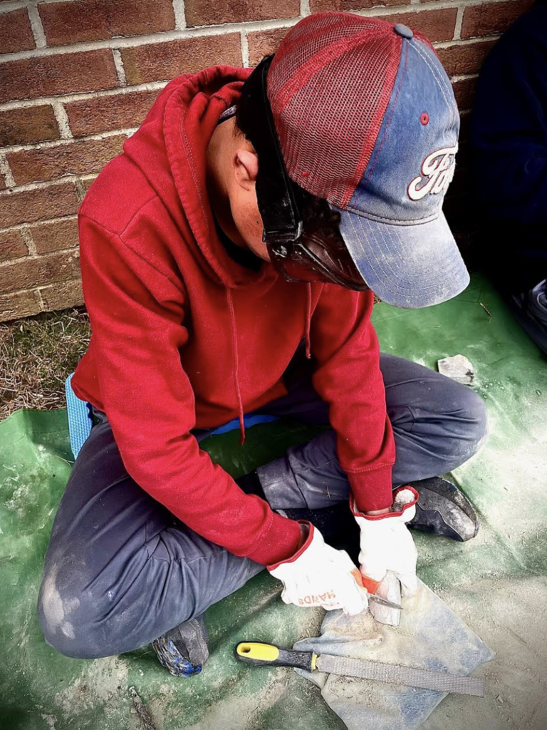 A student wearing a red hoodie, gloves, and protective eyewear uses a metal tool to carve a piece of stone while sitting on a mat outdoors near a brick wall.