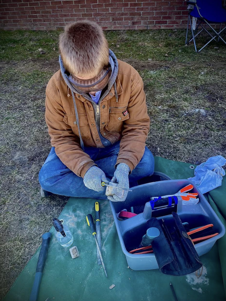 A student wearing a brown jacket, gloves, and safety goggles carefully files or shapes a small piece of stone while sitting cross-legged on a mat, with carving tools nearby.