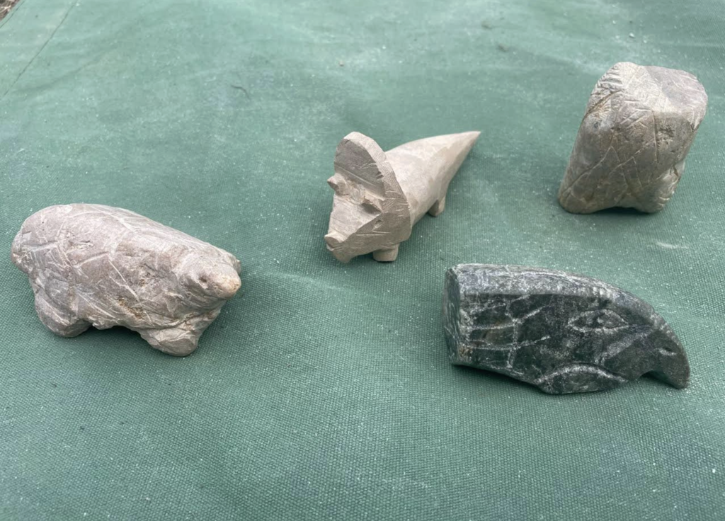 Four small soapstone animal carvings displayed on a green surface, including a bear-like figure, a stylized animal with a pointed tail, and other abstract animal shapes.