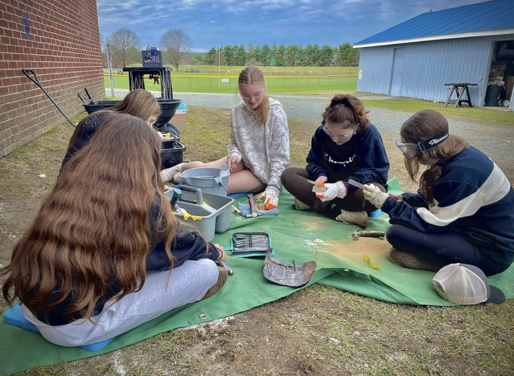 A group of students seated in a circle on outdoor mats work together on stone carving projects, using hand tools and wearing safety gear, with school buildings and a field in the background.