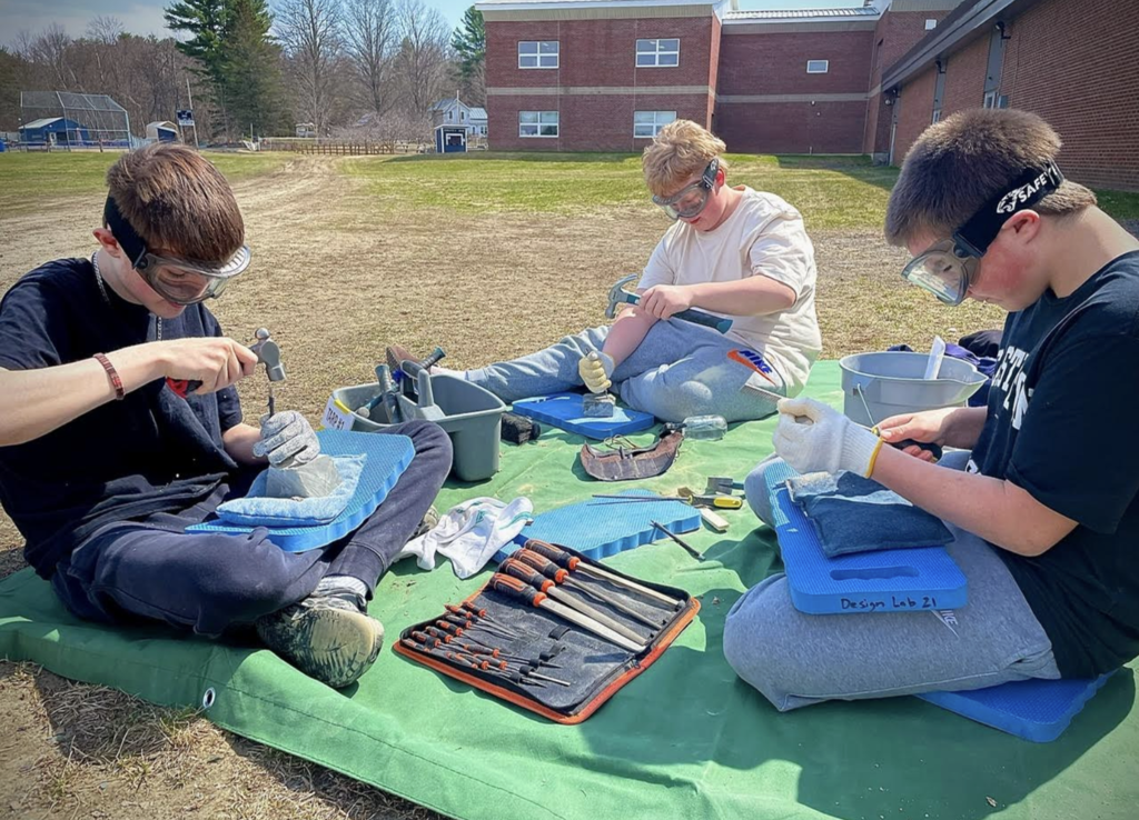 Three students sitting outdoors on mats wearing safety goggles and gloves use hammers and carving tools to shape soapstone pieces, with toolkits and materials spread out in front of them.