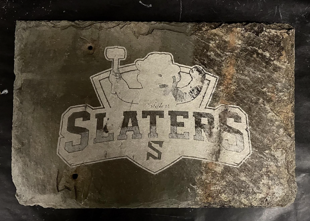 A rectangular slate tile engraved with the word “SLATERS” and an illustration of a worker holding a hammer, showing contrast between etched and natural stone areas.