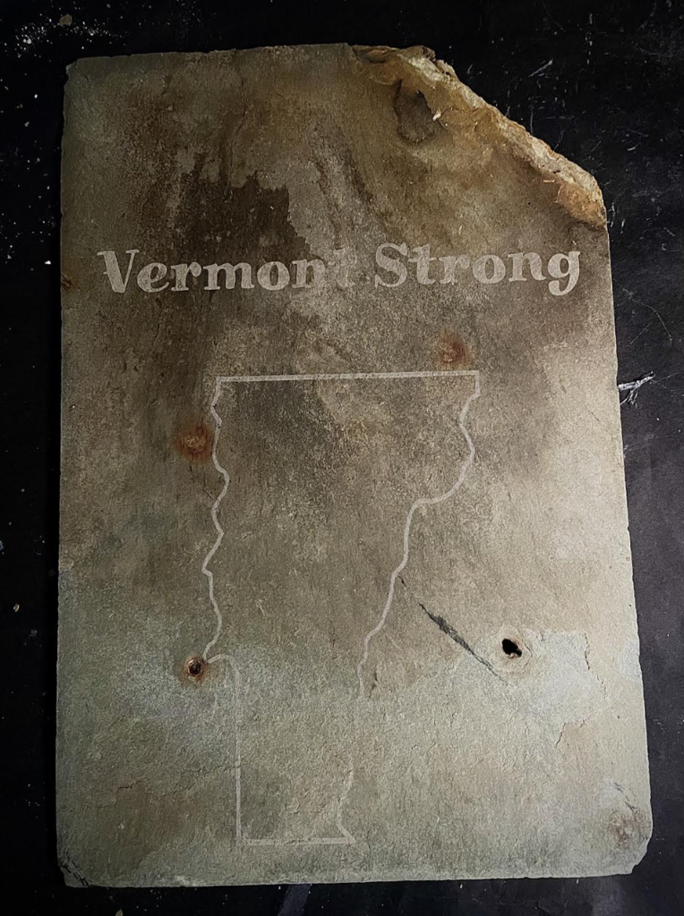 A slate tile engraved with the words “Vermont Strong” above an outline of the state of Vermont, with natural stone texture visible throughout.