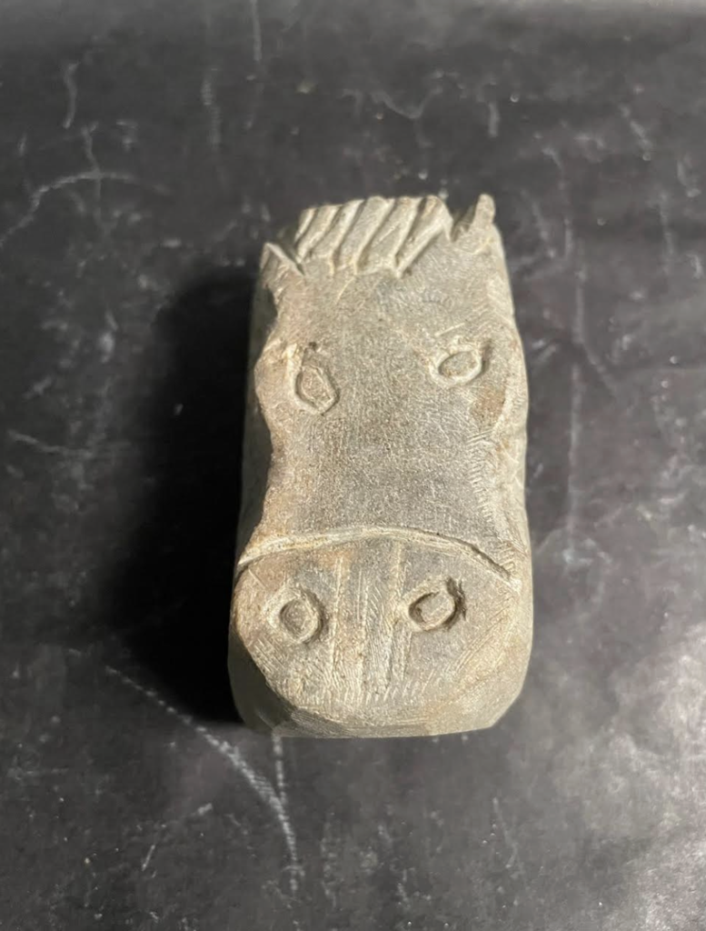 A close-up of a small hand-carved soapstone sculpture shaped like a horse’s head, with simple engraved details for eyes, nostrils, and mane, resting on a dark surface.