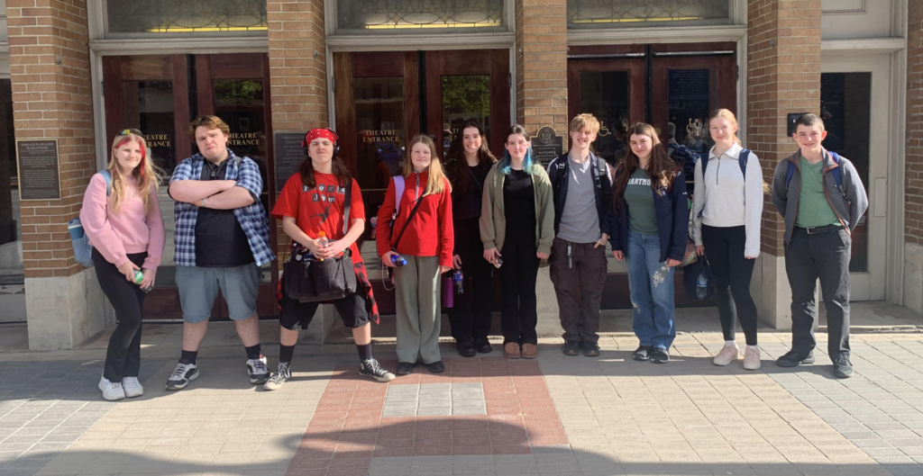 A group of about eleven high school students stand in a line outside the entrance of the Paramount Theatre on a sunny day. The students are casually dressed, some wearing backpacks and holding water bottles, and are smiling or posing for the photo. Behind them are brick walls and glass doors with signage indicating the theatre entrance.