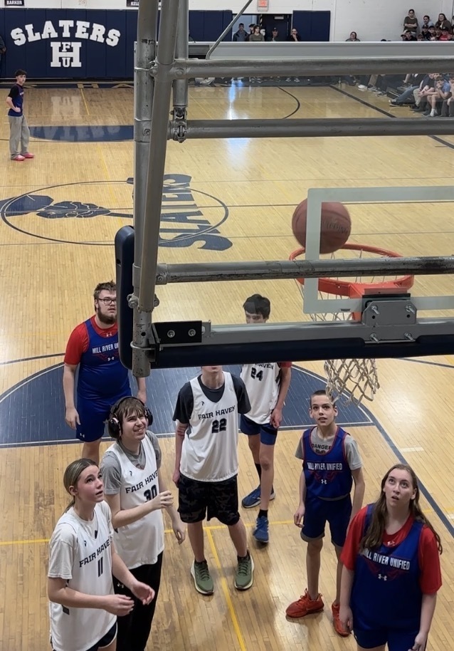 Players from both teams stand under the basket watching the ball as it approaches the hoop, ready for a rebound.
