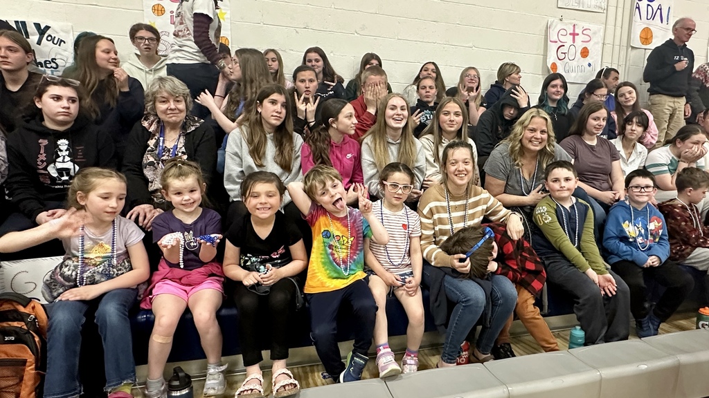 A group of younger students and adults sit together in the bleachers, smiling and interacting, with several children wearing bead necklaces.