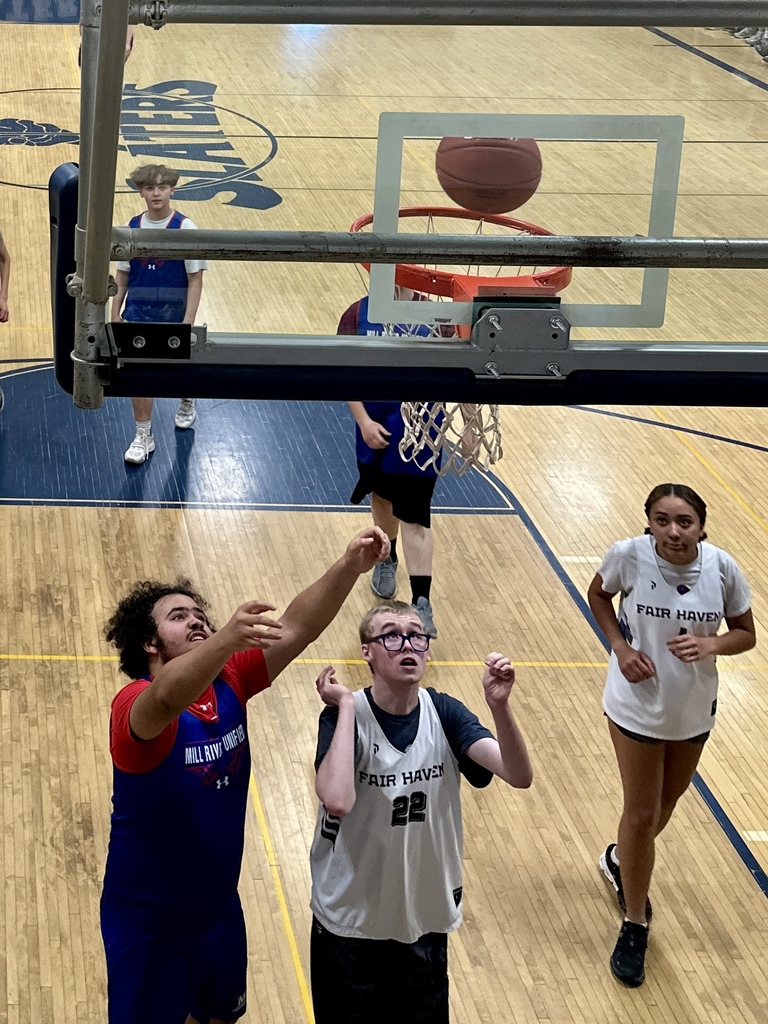 A basketball is captured mid-air above the hoop as players from both teams look up, anticipating the rebound. The backboard and rim are in the foreground.
