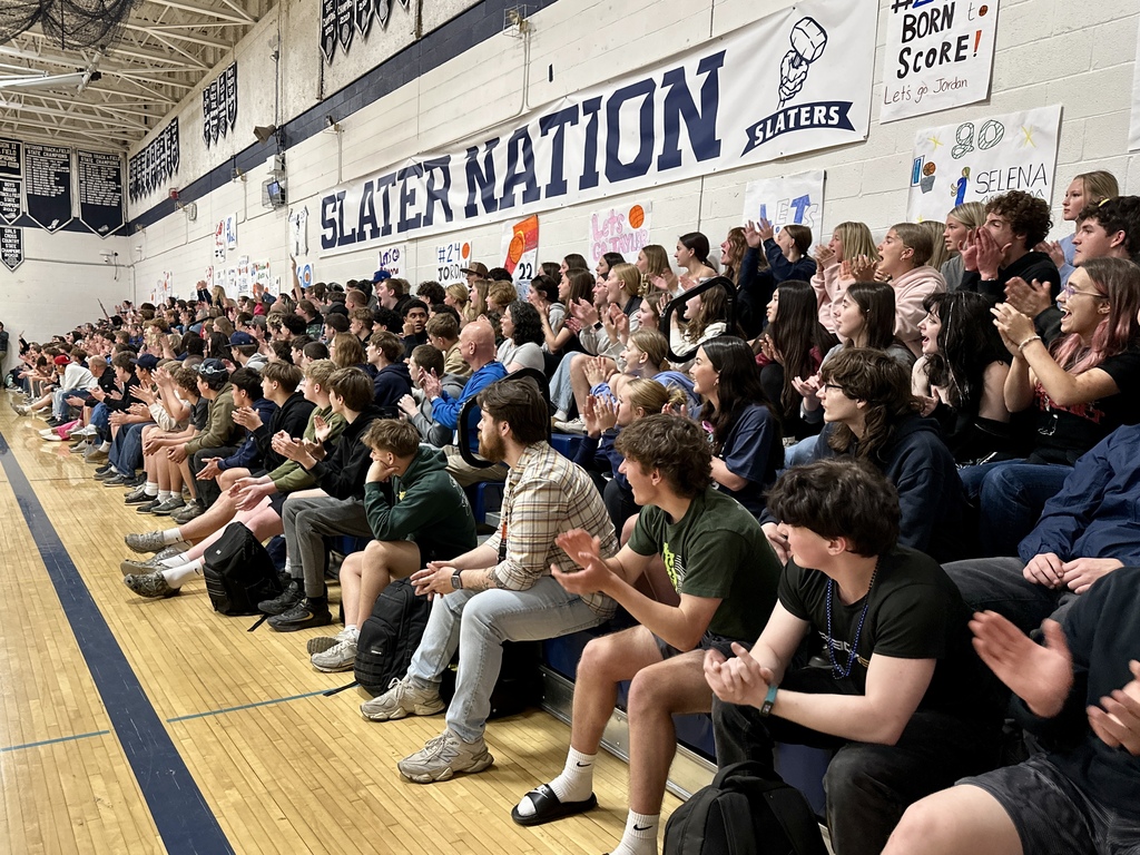 A large crowd of students and staff sit in the bleachers clapping and cheering. A “Slater Nation” banner hangs on the wall above them along with handmade signs.