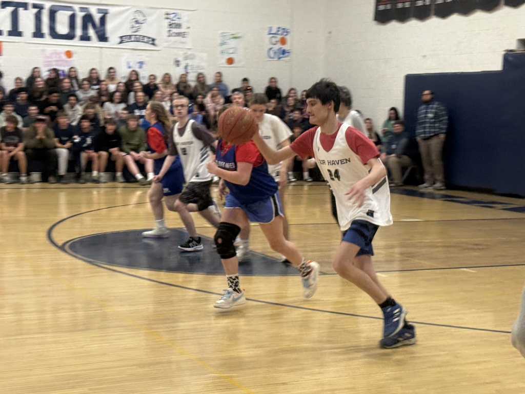 Two players run down the court during a game, one dribbling a basketball while others move into position. The gym is full of spectators watching from the bleachers.