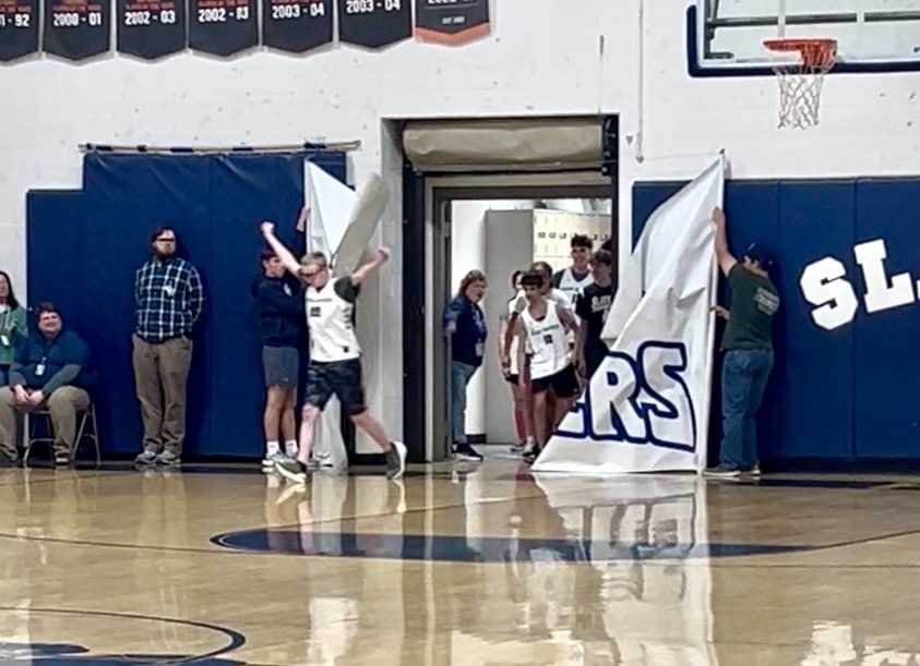 A player runs onto the court through a banner held open by others, raising their arms as teammates and staff stand nearby.
