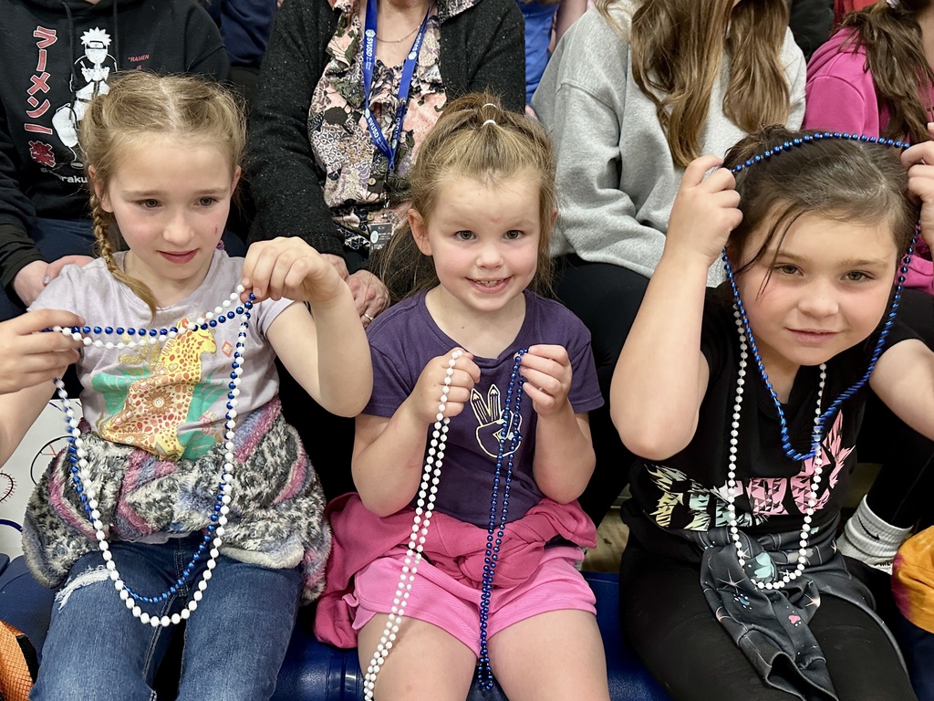 Three young elementary school students sit together in the bleachers smiling and holding blue and white bead necklaces as they watch the game.