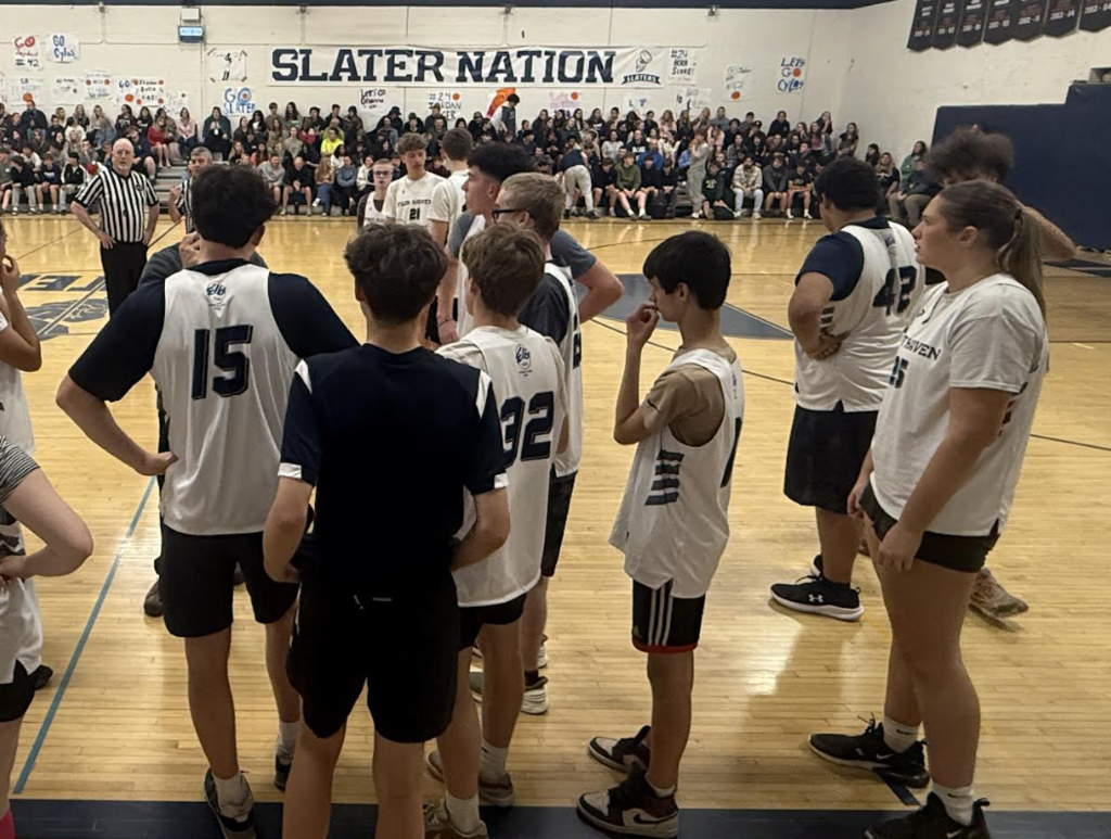 A wider view of the team huddle shows players listening to their coach near the sideline, with a referee and a large crowd of spectators seated in the bleachers behind them.