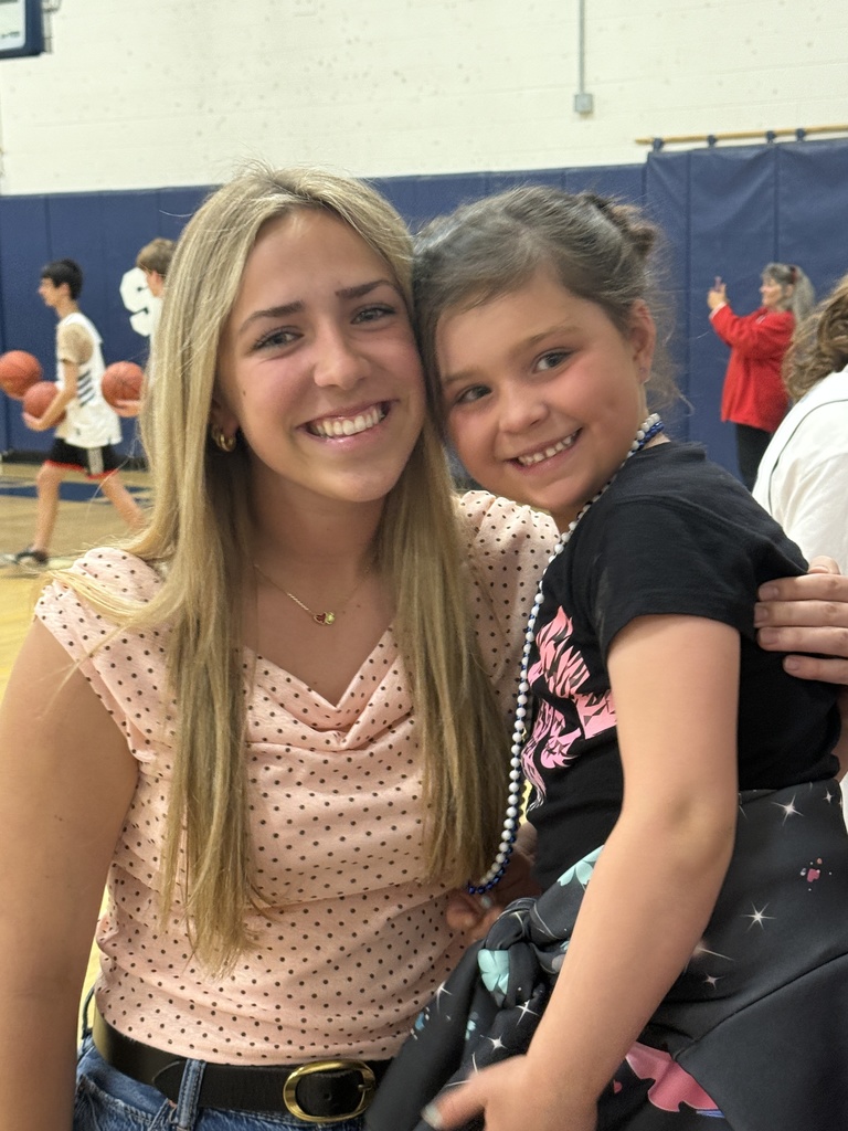 A high school student with long blonde hair smiles while holding a young child wearing beads. They pose together in a gymnasium with basketball players and spectators in the background.