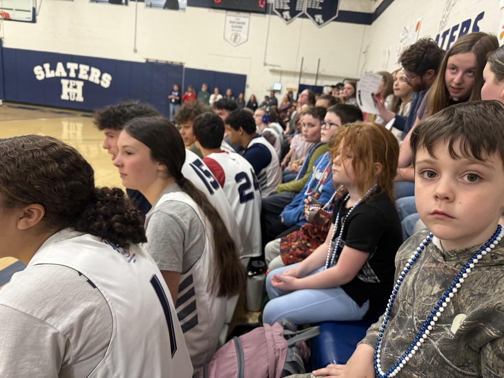 A group of students and young children sit along the bleachers during a game. A young boy wearing blue and white beads looks toward the camera while older students in basketball jerseys sit nearby, focused on the game.