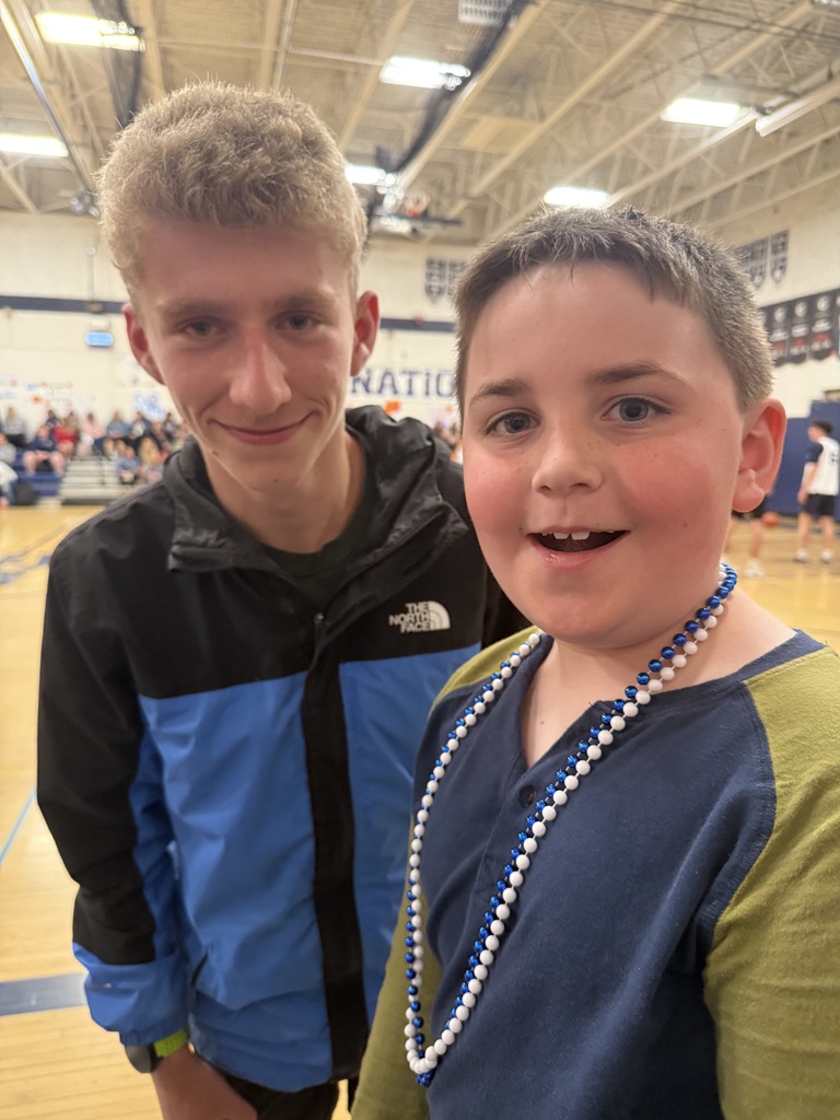 A young student wearing blue and white beads smiles at the camera while standing beside an older student in a blue and black jacket inside a school gym. The basketball court and spectators are visible in the background.